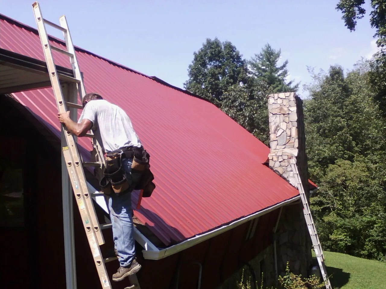 Roofer on a ladder beside a red metal roof and stone chimney — Soffit & Fascia Repair by Roofing Brothers in Massillon